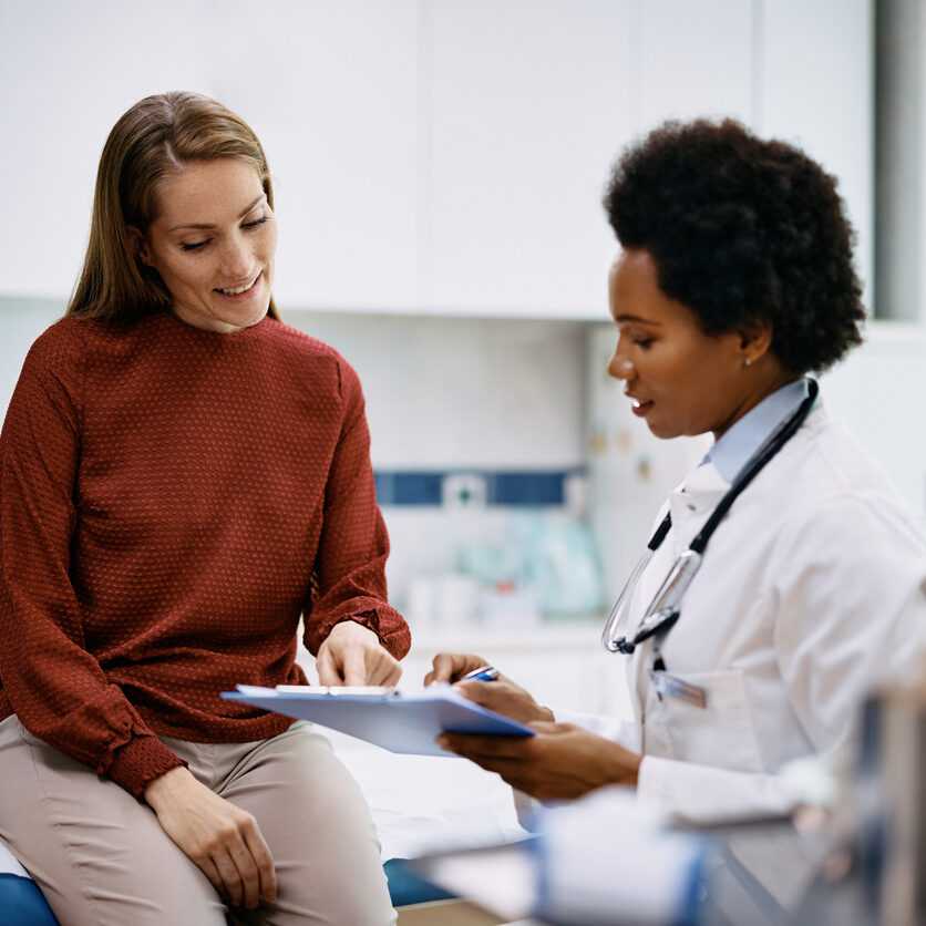 Happy Woman Going Through Her Medical Data With Black Female Doctor At The Clinic.